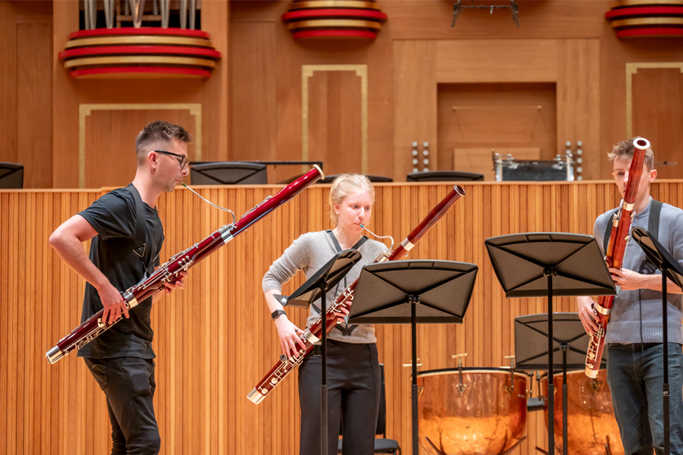 A group of students, holding bassoons, performing in a wind quartet on stage, with wooden panelling behind them.